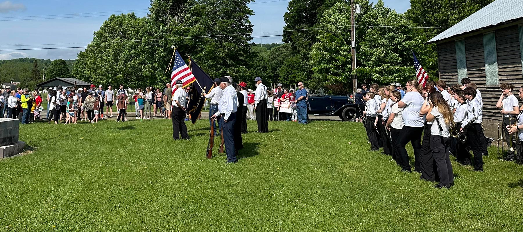 Outdoors in a park with green grass and trees, a group of veterans in uniform, of of whom holds the American flag.